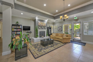 Interior Clubhouse, Couch, sofa chair and coffee table arranged around fireplace, tv on left side of fireplace, bookshelf left of tv, potted plants arranged around room, gray walls, tile floor, exit to pool area behind couch.