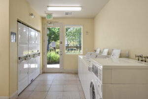 Interior Laundry Facilities, at least 6 dryers and 5 washers, window near entrance door, tile floor, light yellow wall paint.